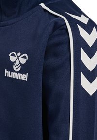 Veste de sport bleu marine en tissu texturé, avec un logo blanc et un motif en chevrons sur les manches, agrémentée de finitions blanches contrastantes.
