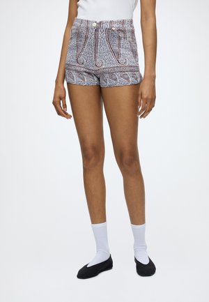 Frau trägt blaue und weiße High-Waist-Shorts mit Paisley-Muster, weiße Socken, schwarze Ballettschuhe und ein weißes ärmelloses Oberteil, steht.