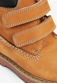 Chaussure en cuir marron avec deux bandes Velcro, accent noir contrastant, couture blanche et finition texturée ; semelle en caoutchouc de couleur brune.