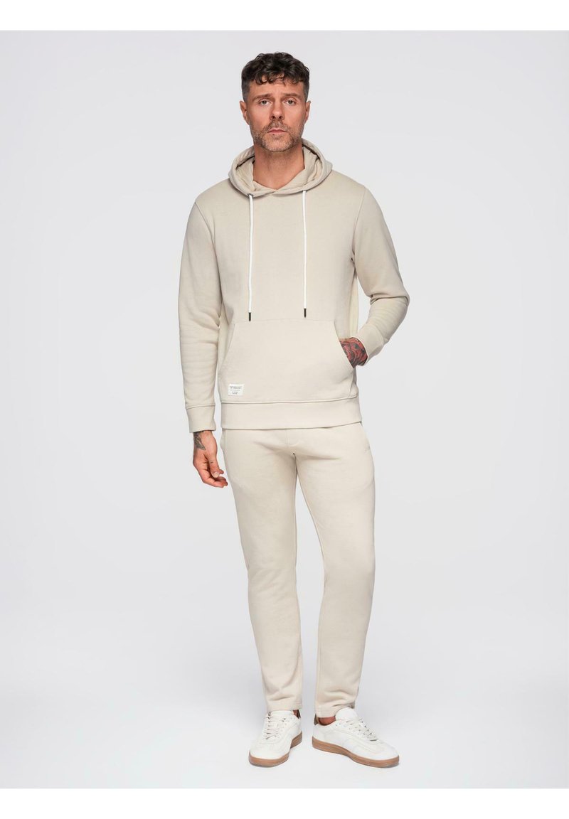 Homme aux cheveux courts et bouclés, tatoué, portant un sweat à capuche beige et un pantalon de survêtement assorti, debout avec une main dans la poche et des baskets blanches.