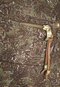 Bruine stof met groen paisley-patroon, voorzien van een gouden ritssluiting en leren trekker. Gestructureerd oppervlak met zichtbare stiksel details.