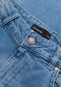 Jean en denim bleu clair avec un bouton en métal argenté et une étiquette de marque noire portant le texte "ombre". La texture est lisse avec des motifs légers.