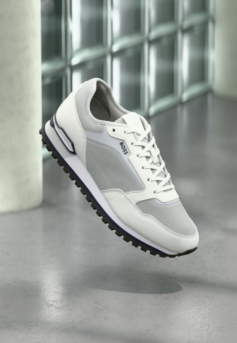 Chaussure de sport blanche avec tige en mesh et cuir, accents gris, lacets, semelle extérieure en caoutchouc rainurée, et un logo sur le côté.