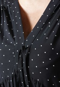 Zwarte blouse met witte stippen, V-hals, geplooide stof bij de schouders en een strikdetail bij de halslijn. Soepele textuur.