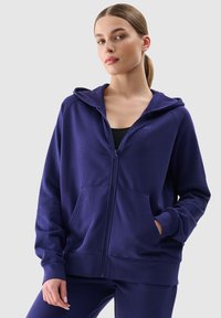 Felpa zip con cappuccio in tessuto viola scuro, dotata di cappuccio, tasche frontali e polsini a costine. Texture liscia con un discreto dettaglio del logo.