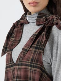 Karirana predpasnik v burgundski, črni in zeleni barvi, z metuljčki na rami. Siva turtleneck majica spodaj, z globokim V-izrezom. Mehka tekstura tkanine.