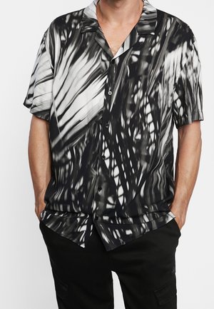 Mand iført sort-hvid abstrakt print kortærmet skjorte med krave, hænderne i lommerne på sorte bukser.