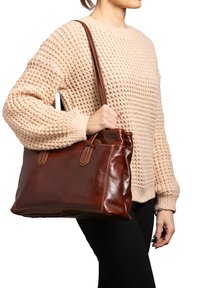 Borsa a tracolla in pelle marrone con doppi manici, texture liscia e dettagli di cucito, tenuta da una persona che indossa un maglione lavorato a maglia beige.