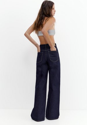 Vrouw met lang haar die een grijze buis-top en hoge taille, wijd uitlopende donkerblauwe spijkerbroek draagt, met haar handen in de achterzakken, zijwaarts kijkend.