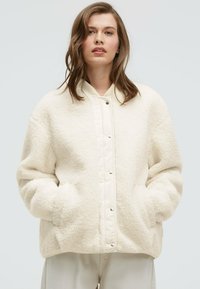 Cream fleece jacket with a textured finish, snap button front, and ribbed collar. Features side pockets and an oversized fit.