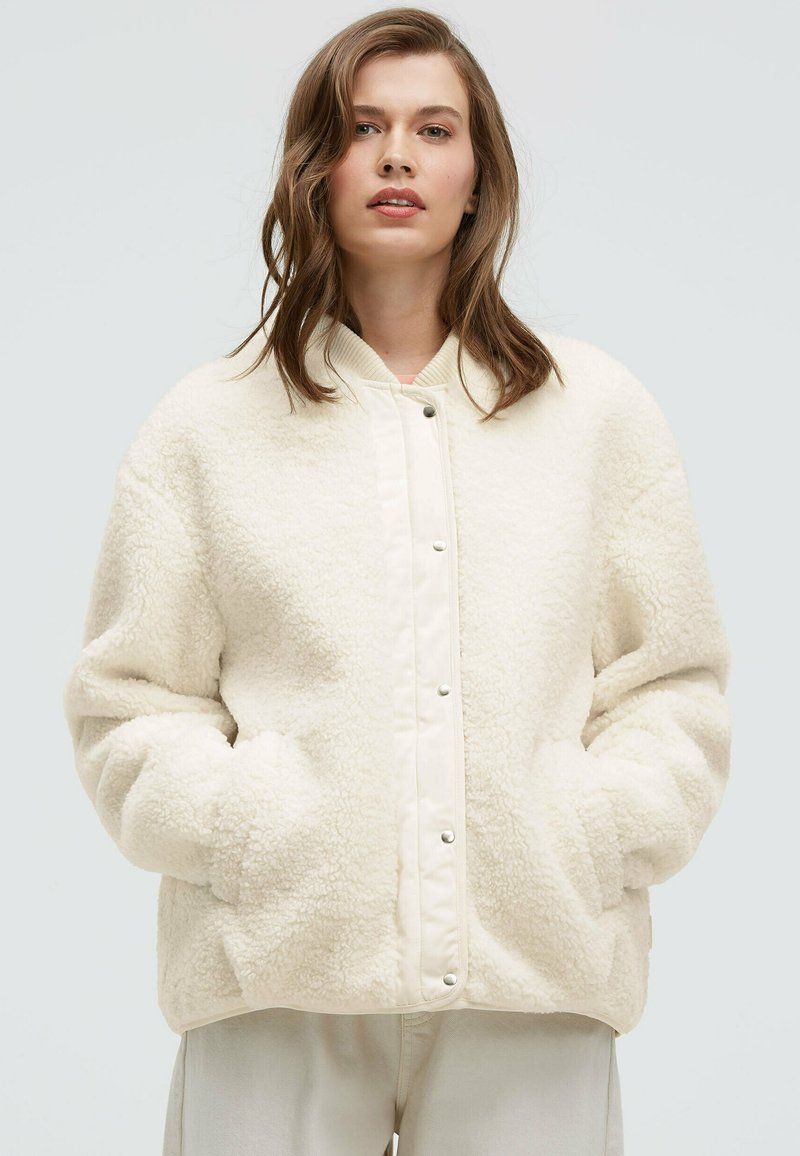 Cream fleece jacket with a textured finish, snap button front, and ribbed collar. Features side pockets and an oversized fit.
