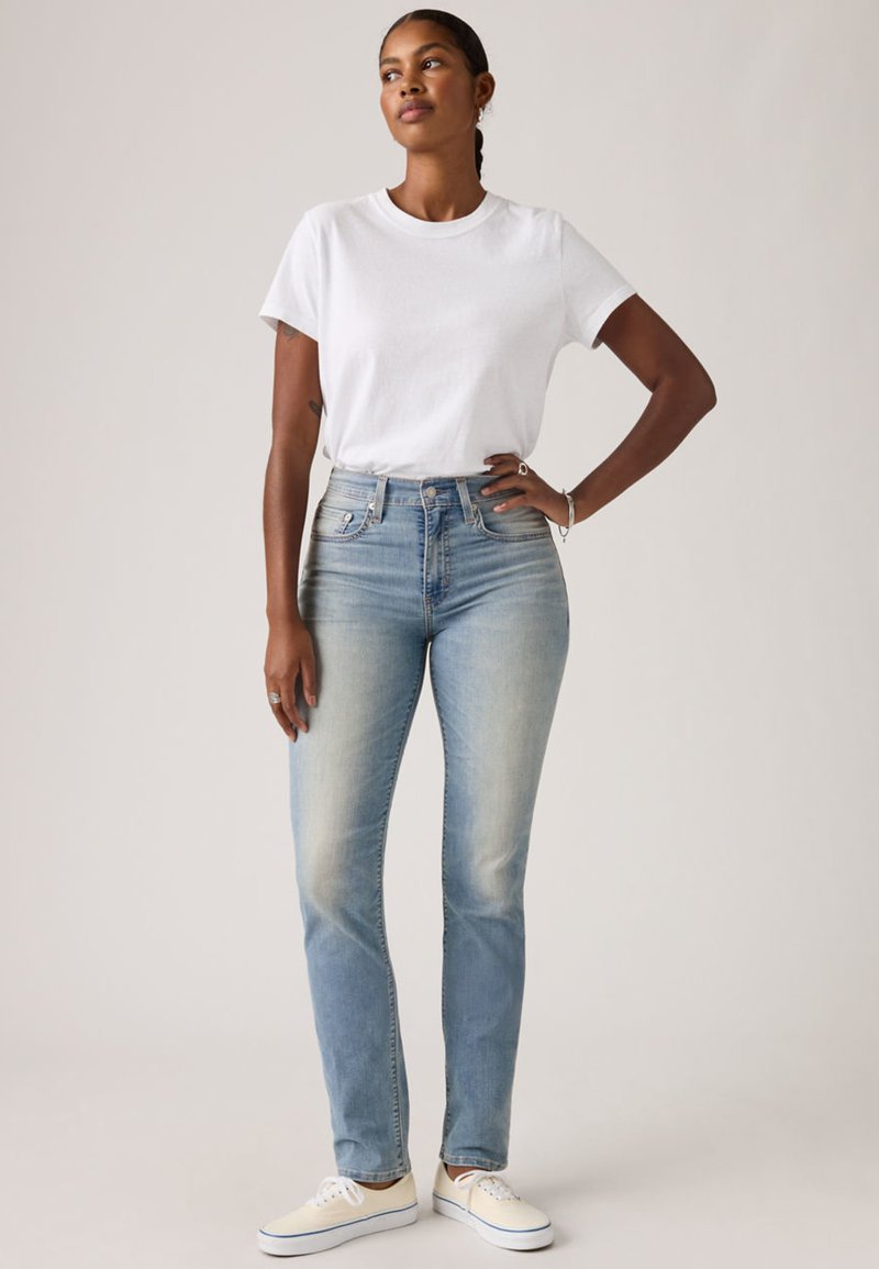 Femme à la peau foncée portant un t-shirt blanc, un jean taille haute bleu clair et des baskets blanches, debout avec une main sur la hanche.