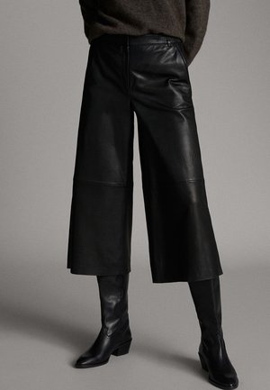 Pantalon en cuir noir à jambes larges et cropped, associé à des bottes en cuir noires montant jusqu'aux genoux, sur un fond neutre.