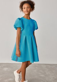 Bla dresses in hellblau mit einem strukturierten Logo-Muster, einem ausgestellten Rock und seitlichen Streifen in Dunkelblau und Weiß. Kombiniert mit weißen Turnschuhen.
