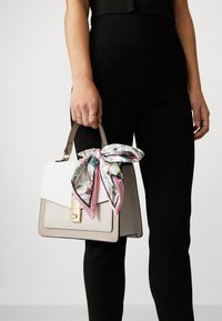 Sac à main beige avec un rabat texturé blanc, des accessoires en or, et une écharpe en soie à motif floral nouée sur la poignée. Matériau lisse, forme structurée.