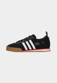 Μαύρο αθλητικό παπούτσι Adidas Samba με λευκές ρίγες, λαστιχένια σόλα, κόκκινη λεπτομέρεια και χρυσό λογότυπο "adidas Samba" στο πλάι, φαίνεται από την εξωτερική πλευρά.