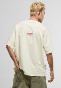 Cremefarbenes, übergroßes T-Shirt mit kurzen Ärmeln. Auf der Rückseite befindet sich ein roter Logo-Text. Weicher Stoff mit einem lässigen Schnitt.