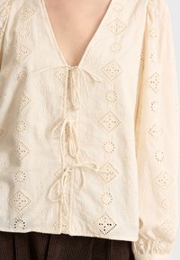 Weiße Baumwollbluse mit tiefem V-Ausschnitt, dekorativer Stickerei und Bindebändern an der Vorderseite, mit langen, puffigen Ärmeln.