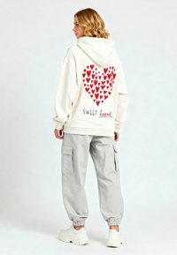 Felpa cremosa oversize con motivo a cuore rosso nella parte posteriore e testo "SWEET heart", abbinata a pantaloni cargo grigi chiari e scarpe da ginnastica bianche.