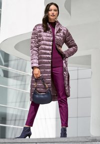 Lange paarse pufferjas met een doorgestikt ontwerp, gedragen over een donkerpaarse top en bijpassende broek. Accessoires omvatten een marineblauwe handtas en blauwe enkellaarsjes.