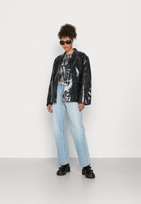 Casaco oversized preto e brilhante com um único botão, combinado com jeans de perna larga azul claro e sapatos pretos robustos.