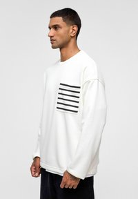 Witte sweatshirt met lange mouwen en een ronde hals. Voorzien van een zwart-wit gestreepte pocket aan de linkerkant. Zachte, gestructureerde stof.