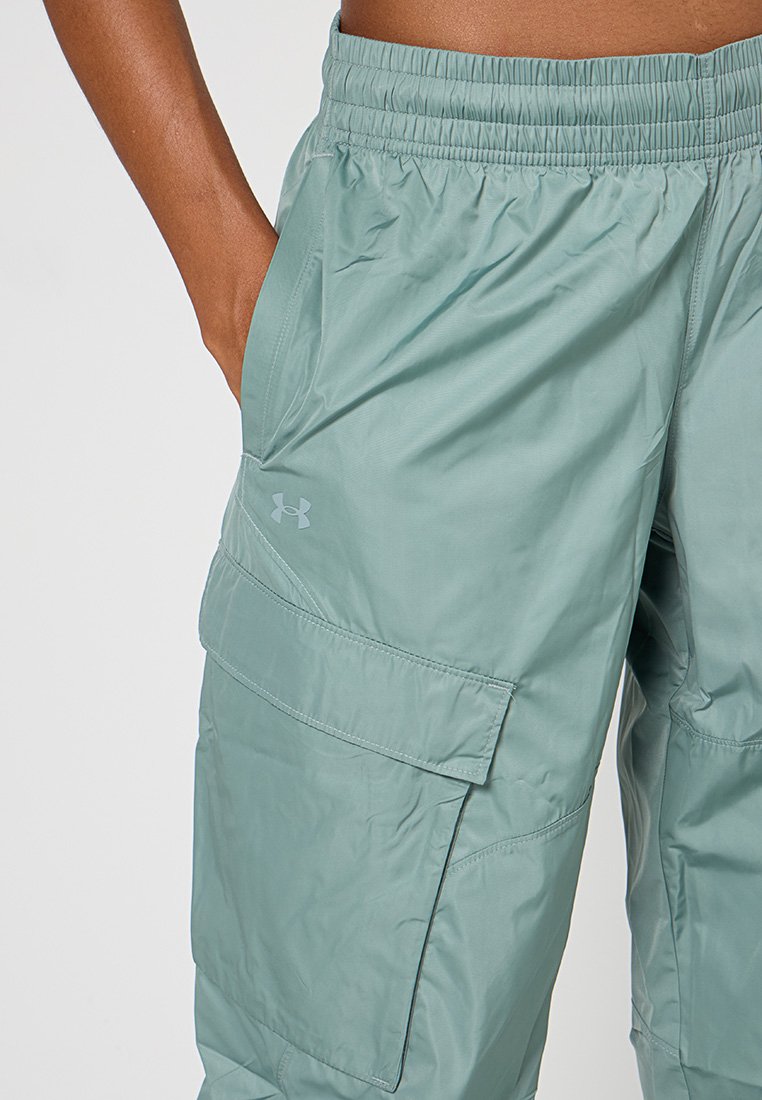 Nahaufnahme von teal-farbenen Cargo-Hosen mit elastischem Bund, Klapptasche und Under Armour-Logo am linken Oberschenkel, Hand in der Tasche.