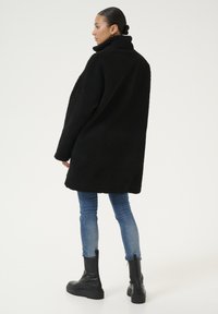 Zwarte nepbontjas met een hoge kraag, losse pasvorm en lange mouwen, gedragen over blauwe skinny jeans en zwarte chunky laarzen.