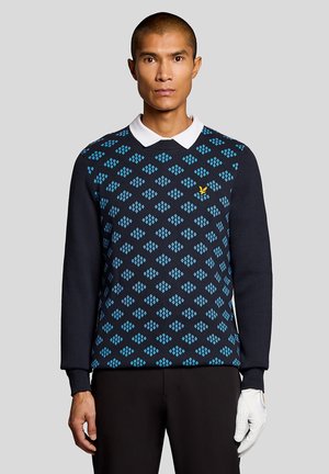 Homme portant un pull bleu marine avec un motif losange bleu clair et un logo jaune, une chemise blanche à col, un pantalon noir et un gant de golf à la main droite.
