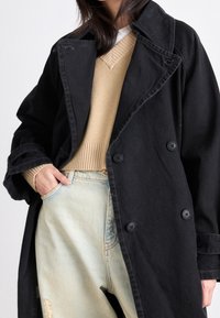 Trench nero doppiopetto in tessuto resistente, con grandi bottoni, vestibilità oversize, abbinato a un maglione lavorato a maglia color cammello e jeans blu.