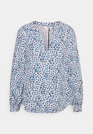 Blouse blanche à manches longues avec motif floral bleu, encolure en V et poignets froncés, présentée sur un fond neutre.
