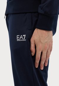 Pantaloni sportivi bleumarin din material moale, cu logo-ul brodat EA7 Emporio Armani pe lateral. Design evazat cu buzunare laterale.