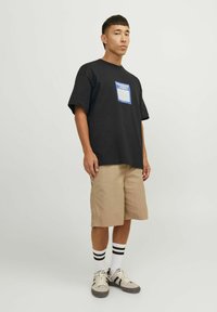 Sort bomulds t-shirt med et blåt grafisk lomme-design, parret med beige shorts, hvide sokker med sorte striber og grå sneakers.