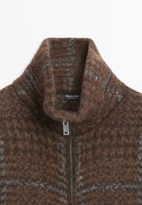 Brązowy sweter z wełny zapinany na zamek, z wysokim kołnierzem, ozdobiony szarą kratką. Materiał ma miękką, teksturowaną powierzchnię.