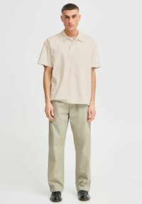 Polo shirt beige à manches courtes avec un col, associé à un pantalon large vert clair. Modèle portant des chaussures noires se tient contre un fond uni.