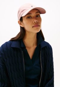 Gorra de béisbol rosa, cárdigan de punto azul marino con patrón texturizado y una camiseta polo oscura. Líneas limpias y contraste de colores suaves.
