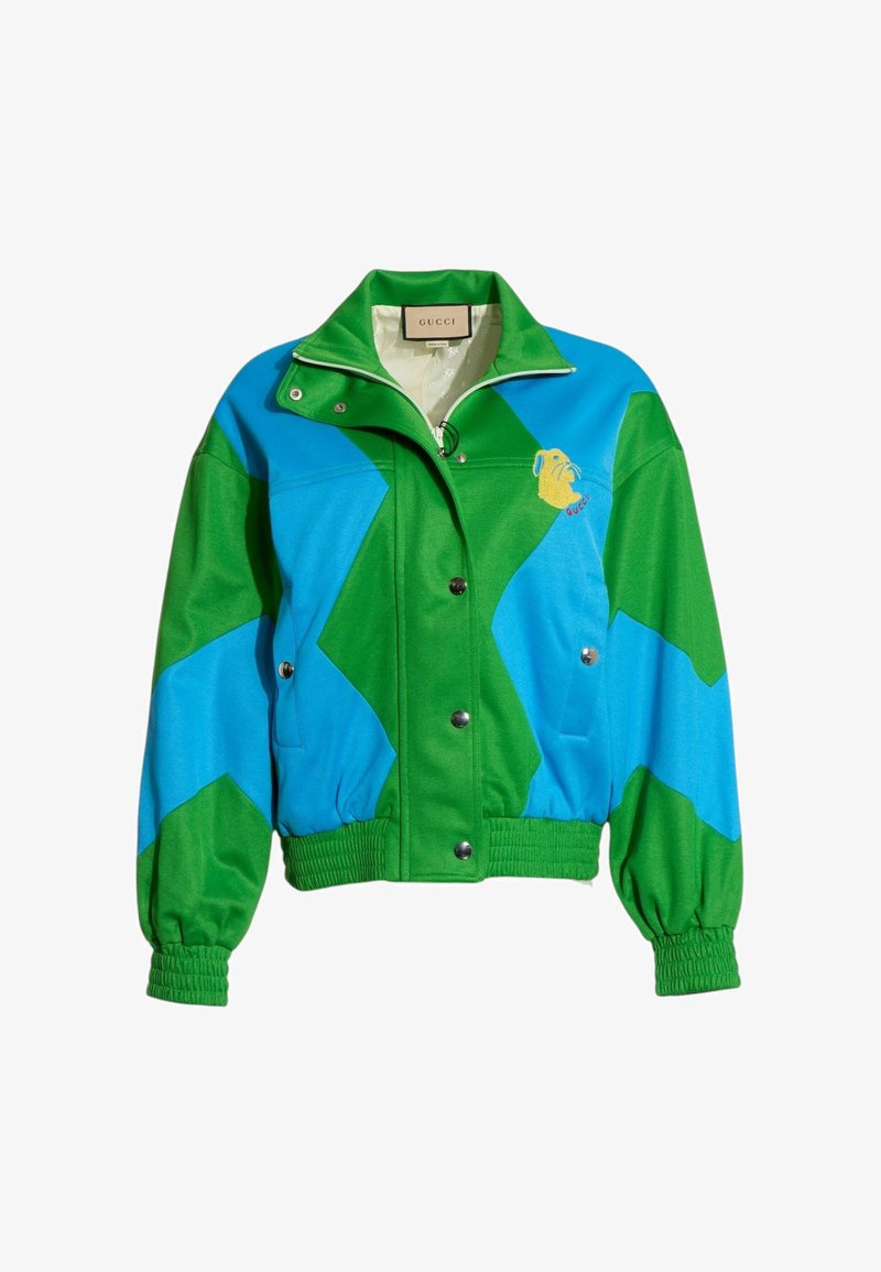 Veste Gucci bicolore verte et bleue avec boutons-pression, poignets et ourlet élastiques, et petit visage d’animal jaune brodé sur la poitrine.