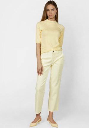 Frau trägt hellgelbes kurzärmeliges Oberteil, hellgelbe gerade geschnittene Hose und dazu passende flache Schuhe vor einfarbig weißem Hintergrund.