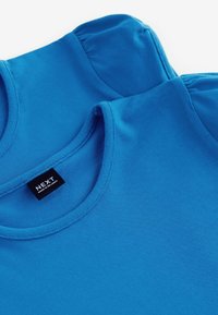 Modré bavlnené tričká s okrúhlym výstrihom a nadúvanými rukávmi. Obsahuje štítok "NEXT" vyšitý čiernou farbou na bielom pozadí.