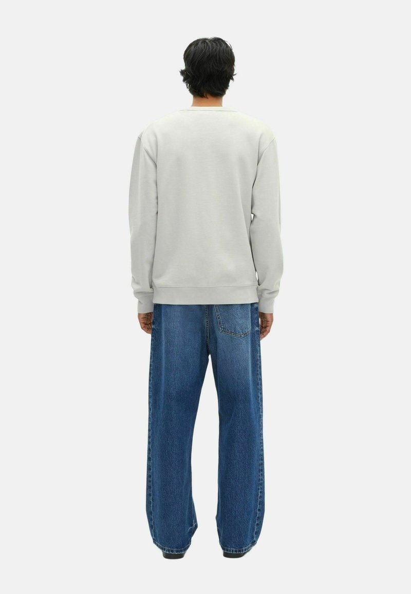 Mann mit dunklen Haaren, der einen hellgrauen Sweatshirt und weite blaue Jeans trägt, steht mit dem Rücken zu einem wei�ßen Hintergrund.