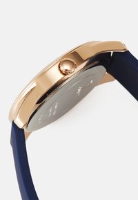 Montre en métal couleur or rose avec un boîtier de forme circulaire, dotée d'un bracelet en silicone bleu et d'une couronne sur le côté. Le dos présente une finition métallisée avec des détails textuels.