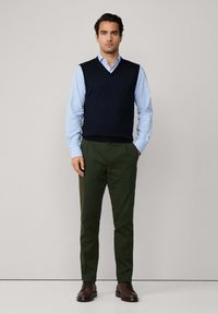 Námornícky sveter s výstrihom do V cez svetlo modrú košeľu, skombinovaný s olivovozelenými nohavicami. Hnedé topánky dotvárajú celkový vzhľad. Jednoduchý, klasický dizajn.