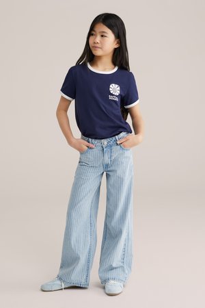 Tüdruk seisab käed taskutes, seljas tumesinist värvi t-särk valge äärisega ja helesiniste triibuliste laia säärega teksadega.