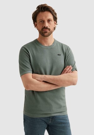 Mann mit kurzen braunen Haaren und Bart, der einen grünen, strukturierten Kurzarmpullover und blaue Jeans trägt, steht mit verschränkten Armen vor einem schlichten Hintergrund.