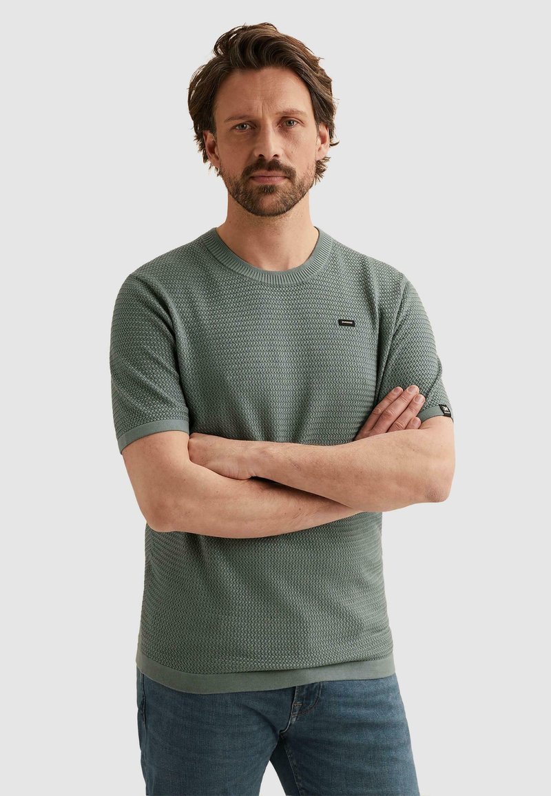 Homme aux cheveux courts châtain et barbe, portant un pull vert texturé à manches courtes et un jean bleu, debout les bras croisés devant un fond uni.
