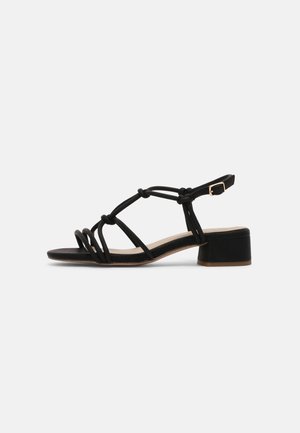 Black low-heeled sandal with knotted straps, open toe, and adjustable buckle ankle strap on neutral background.