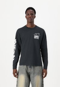 Schwarzes Langarmshirt mit weißem Grafiktext auf der Brust und dem Ärmel, aus Baumwolle mit lässiger Passform, kombiniert mit hellen, distressed Jeans.