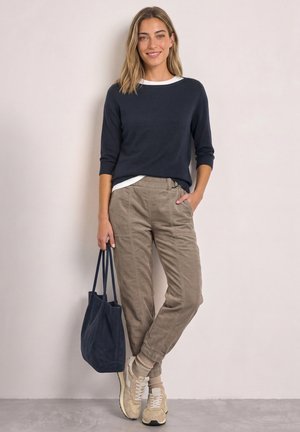 Vrouw staat tegen een muur, draagt een navyblauwe trui, beige ribfluwelen broek, sneakers, houdt een navyblauwe tas vast en lacht naar de camera.