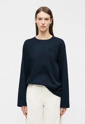 Frau, die ein dunkelblaues Langarmshirt mit Rundhalsausschnitt und dezentem Text auf der Brust sowie weiße Hosen trägt, steht vor einem einfarbigen Hintergrund.