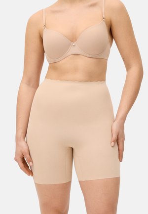 Mujer con pantalones cortos moldeadores beige de cintura alta y sin costuras y un sujetador beige a juego, sobre un fondo blanco.